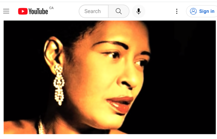 A close up shot of Billie Holiday singing the blues. The lights must be hot. Her face has a sheen. Her lips are red. I never noticed she painted her eyebrows.A screenshot of a YouTube video.
